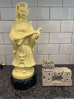 Yellow ceramic Asian figure holding a musical instrument standing on a black base next to the carved stone Foo Dog statue on a countertop with white tiled backsplash.