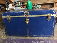 Front view of large blue antique steamer trunk with metal corner guards, latches, and center lock showing wear consistent with age.