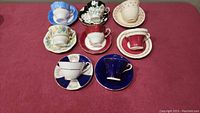 All 8 cups and saucers arranged on a burgundy cloth background showing the variety of colors and patterns in the lot.
