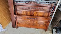 Wooden antique Eastlake style headboard and footboard leaning against a wall showing carved floral and geometric designs with burled wood veneer insets.