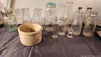 Photo shows a collection of old milk bottle sealers and a cracked butter crock. The crock is ceramic and printed with Dairy Lea Salt Butter. Multiple glass bottles and jars are visible with various sizes and embossed markings.