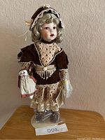 Full view of the porcelain heirloom doll standing on a wooden surface, showing vintage-style dress, lace bonnet, and small fabric bag.