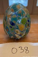 Front view of glass egg paperweight showing colorful embedded millefiori floral patterns on a wooden surface.