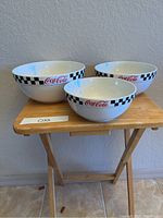 Three white stacking Coca-Cola bowls with checkerboard design on wooden folding table against a beige wall.