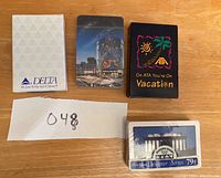 Four decks of playing cards showing the fronts of the boxes on a wooden surface, including Delta Airlines, hotel building night view, ATA Airlines vacation theme, and Las Vegas monument deck
