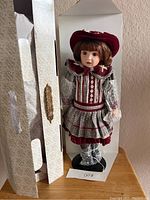 Full view of the doll standing inside its original decorative box, showing the doll dressed in period clothing and displayed on a black stand.