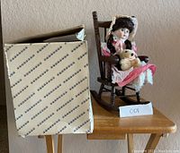 Porcelain doll in pink dress sitting in wooden rocking chair beside original box.