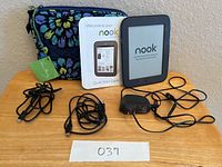 Front view of the Nook eReader powered on with welcome screen, Vera Bradley floral case, USB cable, power adapter, and quick start guide booklet displayed on table.