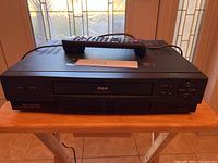 Front view of the black RCA VCR showing the tape slot and basic control buttons with the remote control placed on top.