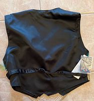 Back side of the black satin vest with tags and small bag of extra sequins attached, showing the tie closure at the back bottom hem.