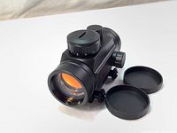 Front angled view of Tasco Propoint 1x30 red-dot sight showing large objective lens with orange tint, brightness adjustment dial (marked 1 to 11), and two lens caps laid out beside the sight.