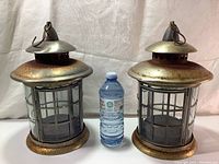 Two vintage large candle lantern holders displayed side by side with a water bottle for scale. Both show a metal rustic body with glass paneling and hanging hook on top.