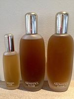 Three bottles of Clinique Aromatics Elixir perfume, two large and one small, placed side by side on carpeted floor against plain white wall.