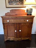 Front view of antique pine cabinet showing single drawer with white knobs and two-door lower cabinet with matching knobs. The top features a scalloped backboard and wood displays weathered finish.