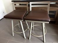 Pair of swivel kitchen counter stools viewed from front, showing brown cushioned seats and metallic frames, each seat about 19x19 inches and 34 inches high.