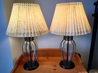 Front view of two table lamps with glass bodies encased in black wire cages and pleated lamp shades placed on a wooden surface.