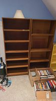 Two tall brown laminate wood grain bookcases side by side with adjustable shelves. Items placed on the floor including electronic cables and boxes of DVDs visible.