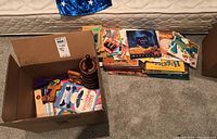 Open cardboard box containing wooden toys and a collection of children's books stacked beside the box on the floor.