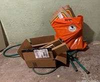 Wide view of pile including cardboard box with wood offcuts, coiled green garden hose, and orange yard waste bags against wall in basement.