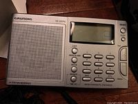 Top view of Grundig YB 300PE radio showing speaker grill, LCD display, and keypad with various function buttons.