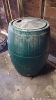 Front angle photo of green plastic rain barrel with lid, showing barrel in garage setting.