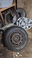 Four Subaru tires mounted on steel rims with visible rust, alongside four white Subaru hubcaps and several grey storage bags, all stored in a garage area with dirt and debris.