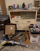 Wide shot of cabinet and items on floor showing tools and supplies including wood tool tray, saw, hammer, paint roller, WD40, and small boxes.