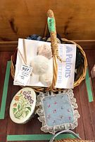 Woven basket containing yarn skeins, knitting pattern book, and two decorative storage tins, placed on the floor.