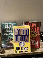 Three books shown upright: 'Shock Wave' and 'Sahara' by Clive Cussler and 'The Road to Omaha' by Robert Ludlum, highlighting some key authors in the collection.