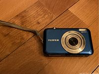 Front view of blue Fujifilm digital camera showing lens and wrist strap on wood surface.