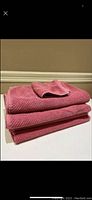 Stack of two full-sized dusty rose SOHO bath towels with a smaller face cloth on top on a white surface against beige wall