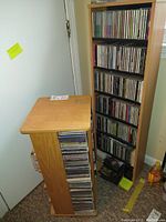 View of two wooden CD storage racks filled with jewel case CDs, one rack taller with black shelves, the other shorter in natural wood finish.