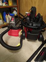 Photo showing two wet/dry vacuums on floor: Craftsman 2-gallon in red/black, Vacmaster with hoses and attachments on top.