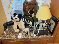Shot of multiple Boston Terrier-themed decor and collectibles on granite counter, including lamp, plush, figurines, mug, and welcome sign showing overall lot content
