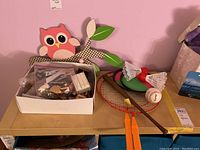 First photo showing dollhouse furniture box, owl and tree branch wall hooks, badminton rackets and shuttlecocks, and a baseball on a shelf.