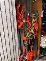 Photo showing multiple extension cords hanging against a metal wall and a wooden cabinet with garden tools including an electric hedge trimmer and lawn trimmer below.