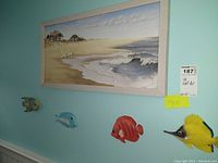 Framed seashore-themed painting showing beach, houses, and ocean waves with four colorful fish wall figures beneath it on light blue wall.