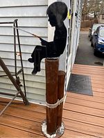 Full view of the metal boy fishing figure sitting on wood piling with rope, displayed on porch with house siding background.
