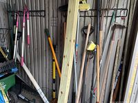 Photo showing a variety of long-handled garden tools including brooms with synthetic bristles, rakes with metal tines, hoes, shovels, and a piece of pressure-treated lumber leaning against a storage wall.