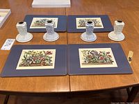 Four floral themed placemats and four white ceramic candle holders arranged on a wooden table.