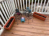 Full view of all items including two brown plastic window planter boxes, black metal bike-shaped planter, two metal seat-shaped planters, painted turtle figurine, and green plastic Ken figure on wooden deck.