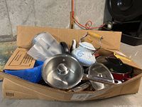 Image of various kitchen cookware items including saucepans, frying pans, teapot, measuring cup and sandwich maker inside a cardboard box.
