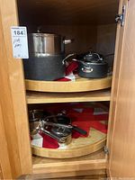Two shelves of cookware inside wooden cabinet showing multiple pots and pans including black granite coated and stainless steel pieces