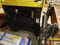 Side and front view of two black plastic sawhorses with yellow top bars and adjustable height markings next to a folding work bench showing folded legs and plastic frame.