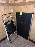 Photo shows two metal step stools with brown frames and black steps stacked next to a large black fabric bulletin board with silver trim in a room with wood paneling.