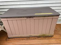 Full view of rectangular plastic outdoor storage chest in beige with brown lid. Shows mildew along bottom edge and dirt on brown deck beneath.