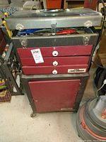 Front view of weighted metal tool cabinet on wheels with three red drawers and hinged top compartment containing assorted tools