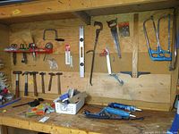 Overall view of wooden pegboard and workbench with various hand tools including hammers, saws, clamps, caulking guns, and other items hung and placed on the bench.