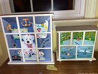 Two painted wooden trinket drawer units on a wooden table by a window. Larger unit with dog and bird painting is approximately 12x12 inches, smaller with aquatic scene is 10x9 inches.