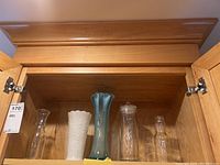 Four vases displayed inside wooden cabinet: clear small glass vase, white milk glass hobnail vase, teal blue art glass vase, clear glass cylinder vase.